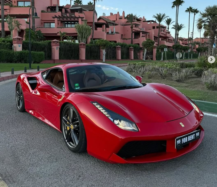 A luxury red Ferrari sports car parked on a road in front of a Mediterranean resort.