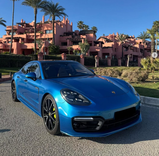 A metallic blue Porsche Panamera parked in front of a luxury pink Mediterranean resort with palm trees.