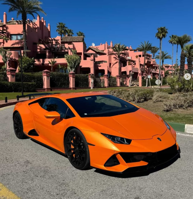 Orange Lamborghini Huracan supercar parked in front of pink Mediterranean villas and palm trees.