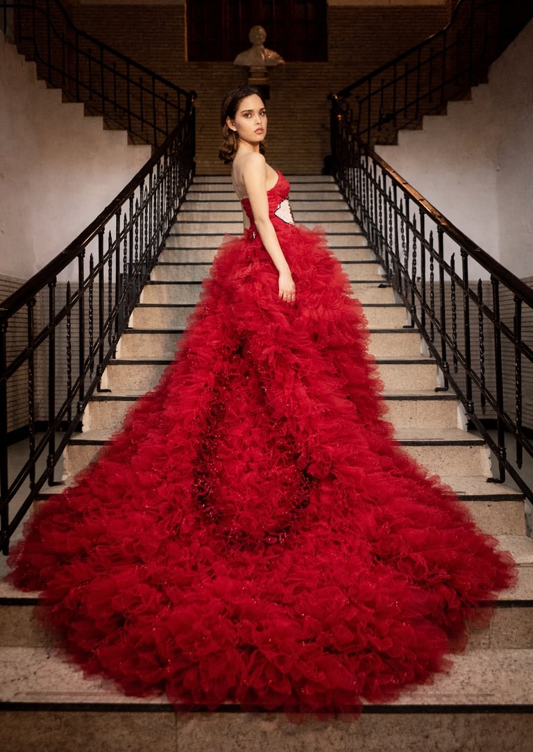 A woman in a voluminous red ruffled ball gown poses on a grand stone staircase.