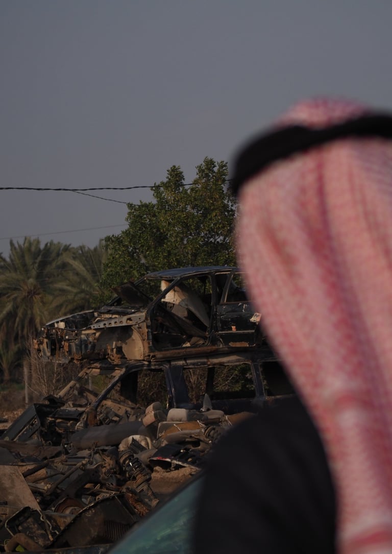 an arab man in a red and white scarf looking at a destroyed car in the by the palm trees in Iraq
