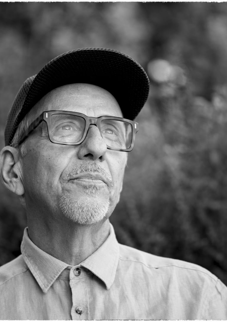 Black and white portrait of a senior man with glasses and a cap looking upwards outdoors.