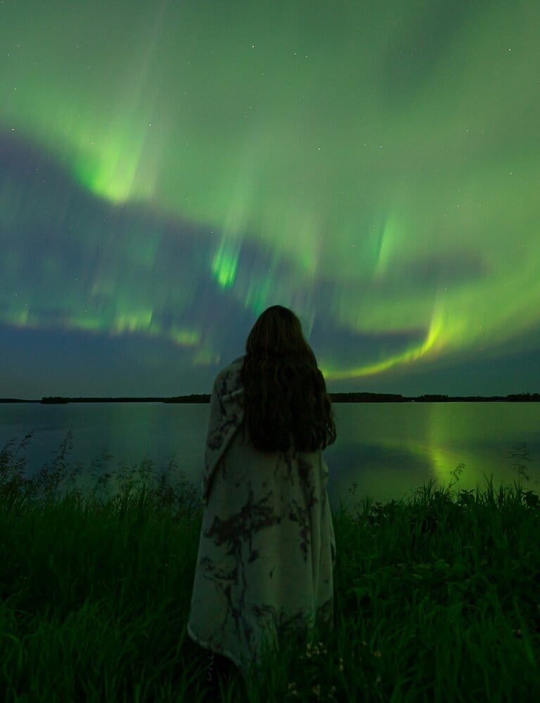 A person watching the vibrant green Aurora Borealis northern lights reflected over a calm lake at night.