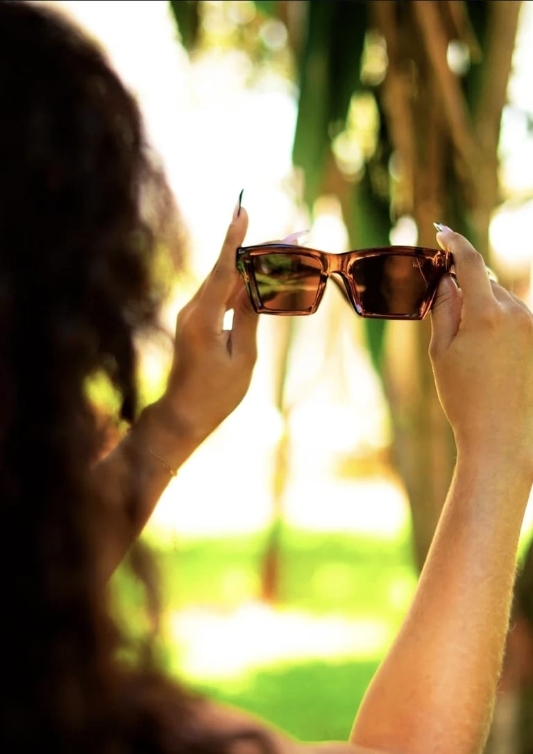 Woman holding up stylish tortoise shell rectangular sunglasses outdoors in bright sunlight.