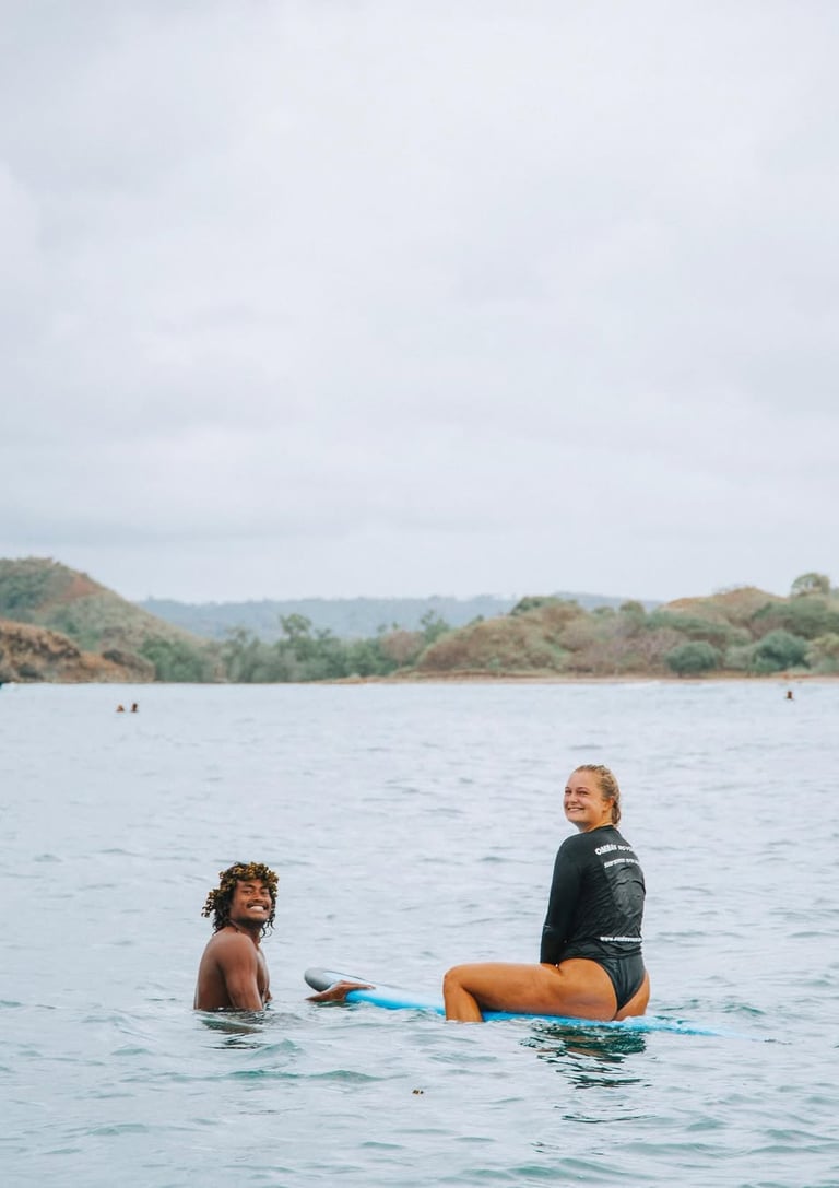 Smiling woman on a blue surfboard and male surf instructor in the ocean with hills in the background.