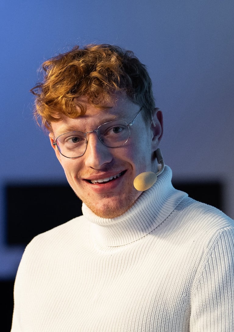 Moderator Przemysław Żuk, kurz Przemek Żuk, moderiert lächelnd eine Paneldiskussion. Er trägt ein Headset-Mikrofon.