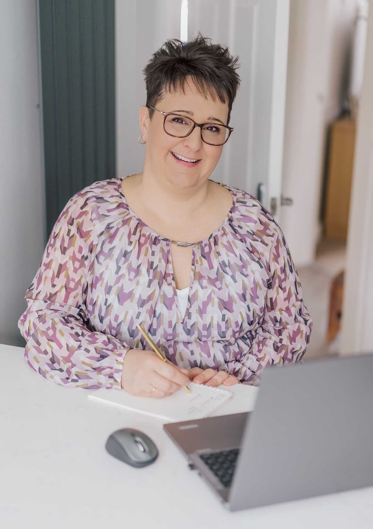 A smiling professional woman with glasses taking notes at her desk with a laptop.