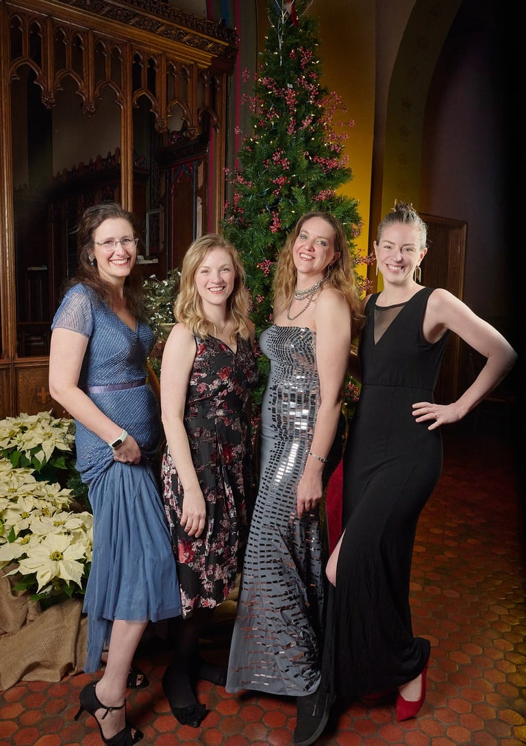 Four women in elegant evening gowns pose together in front of a decorated Christmas tree.