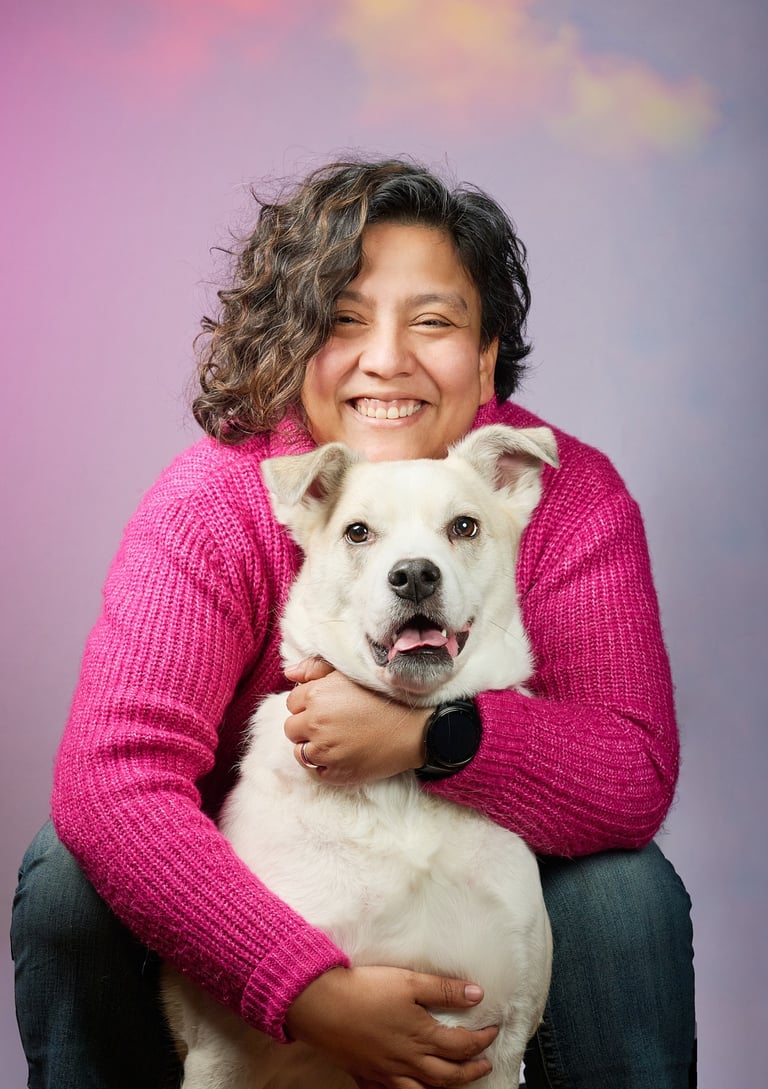 Smiling woman in a pink sweater hugging her large white dog against a purple background.