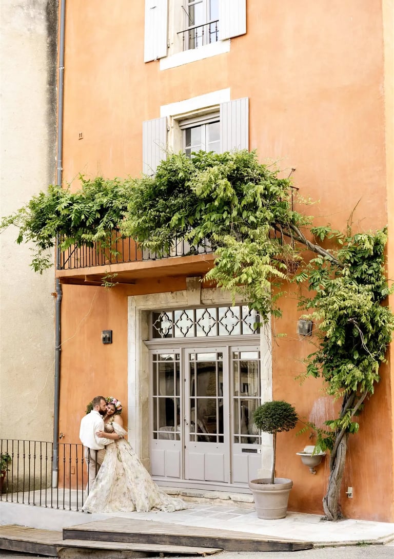 Wide shot of couple in front of ochre building with climbing vine — kiss on the cheek, fine art