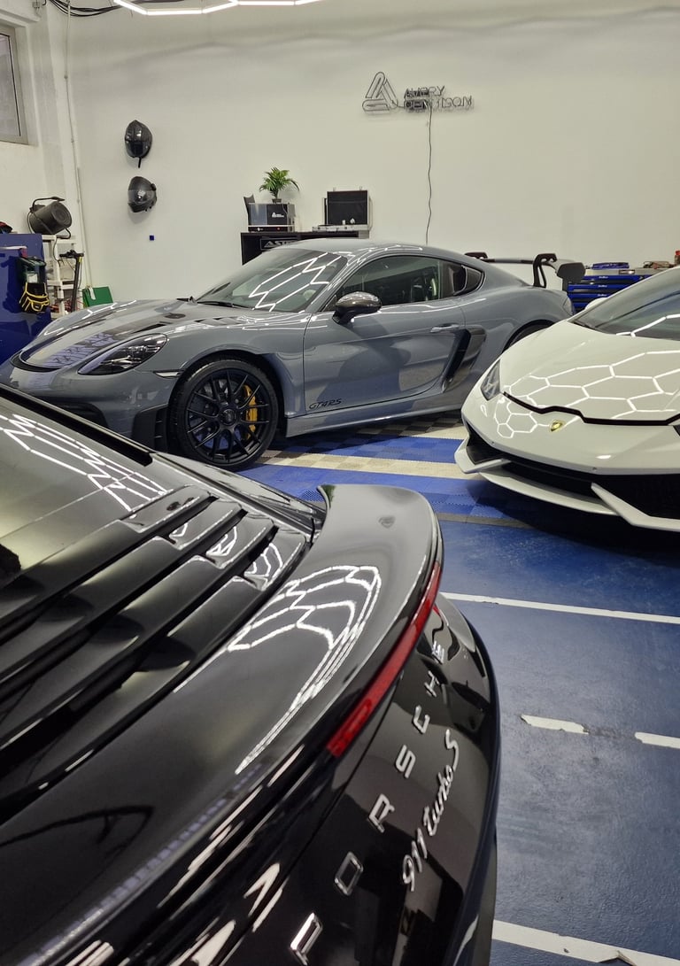 Luxury sports cars including a white Lamborghini and grey Porsche GT4 RS parked inside a modern detailing garage.