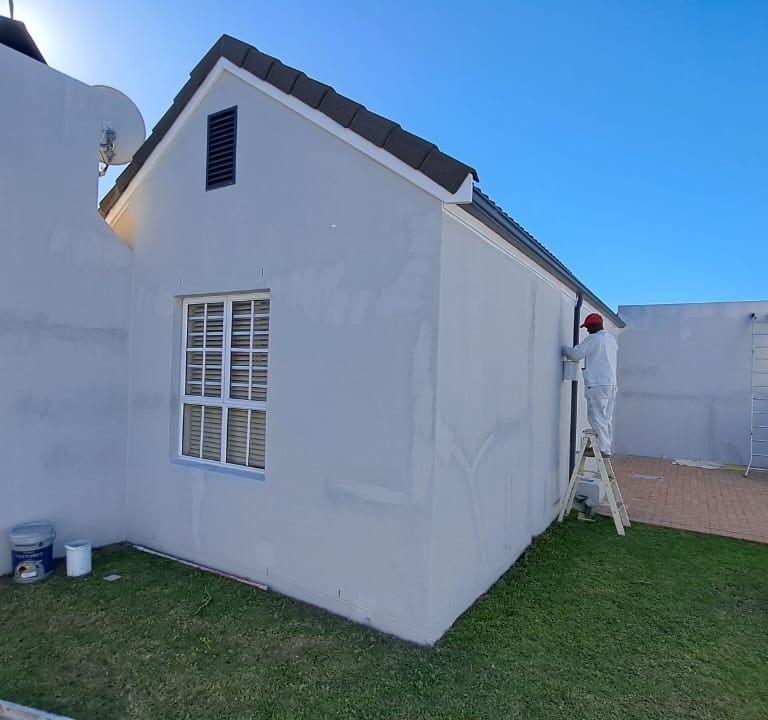 Professional house painter on a ladder applying white paint to a residential home exterior wall.