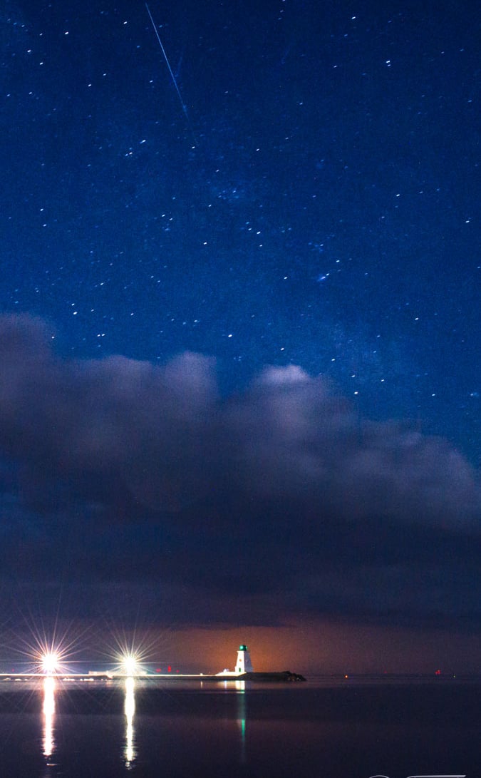 A glowing lighthouse under a starry night sky with a meteor and clouds over the ocean.