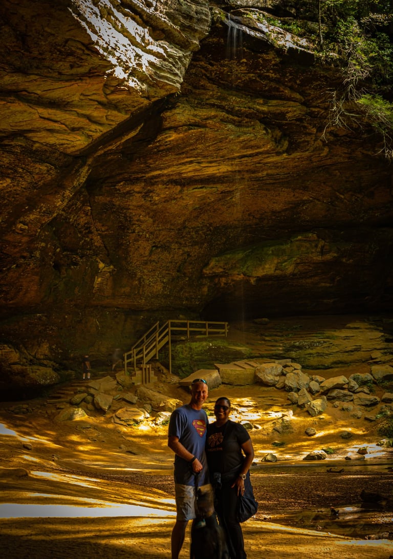 A couple and their black dog stand before a large cave entrance to a small waterfall, Ash Cave Logan