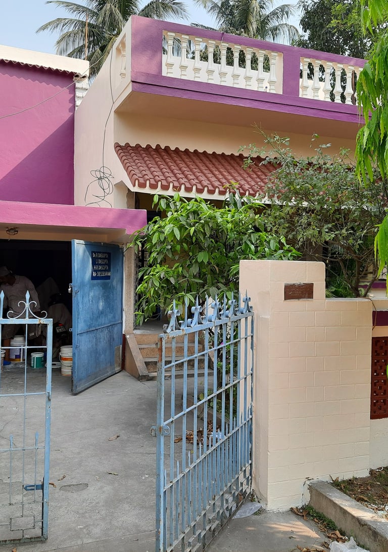 Modern purple two-story house with a blue metal gate and lush green tropical trees.