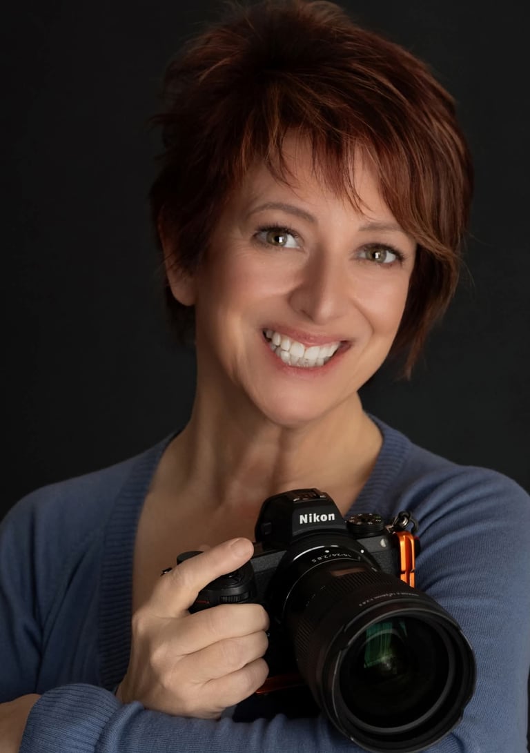 A smiling woman with short auburn hair holding a professional Nikon mirrorless camera.