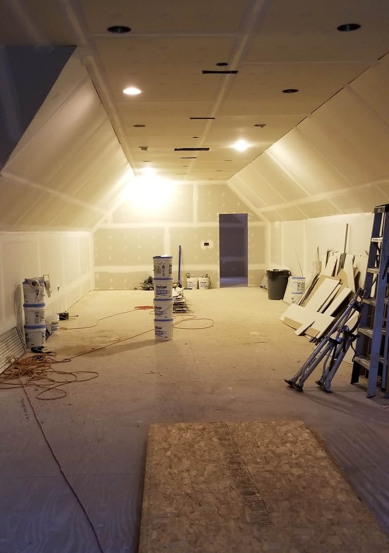 Indoor construction site showing a room with new drywall installation, ceiling lights, and joint compound buckets.