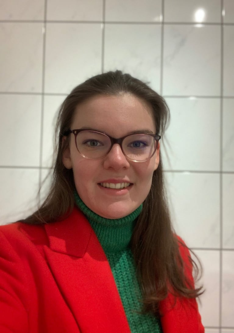 A smiling woman with glasses, wearing a red blazer and green top, standing in a bright room.