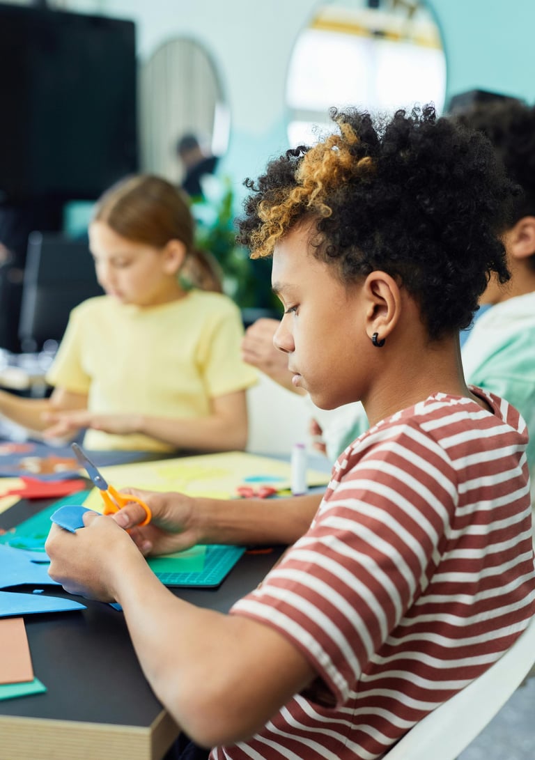Criança com cabelos cacheados demonstrando alta concentração ao utilizar uma tesoura para recortar papel azul
