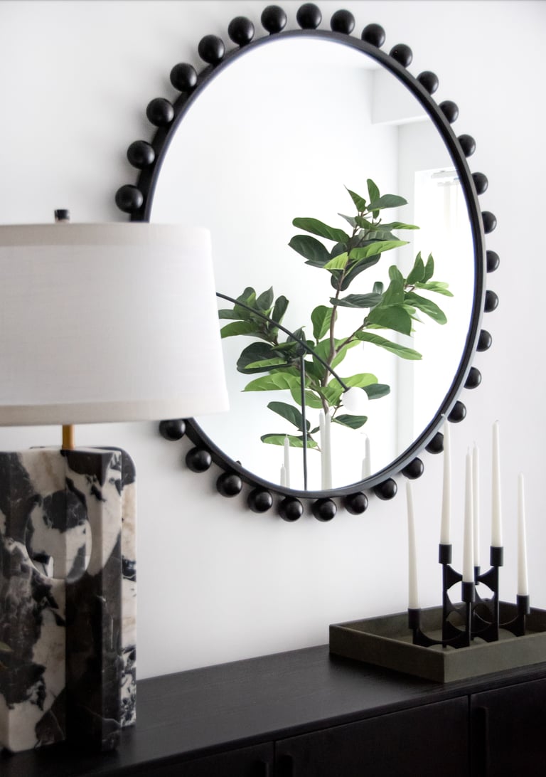 Modern black bead round mirror reflecting a fiddle leaf fig plant next to a marble table lamp.
