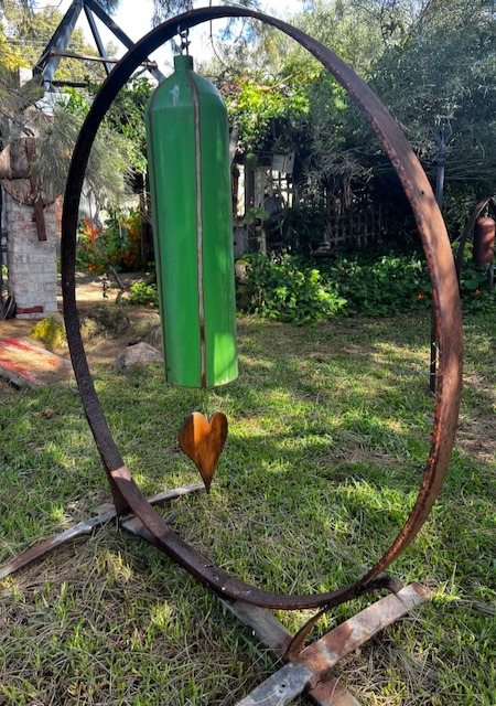 Rustic garden art featuring a large green metal wind chime hanging inside a circular iron frame on a lawn.