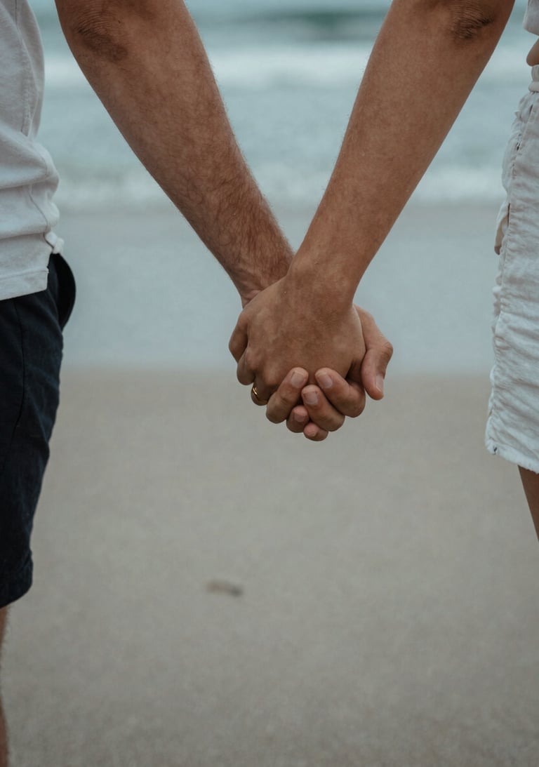 A close-up of a couple holding hands while walking on a North American beach. Soft sand and charcoal blue water in the background. Cinematic, authentic photography.