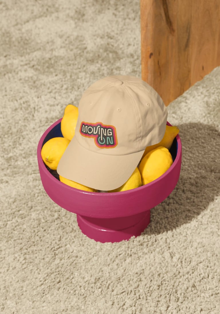 Gorra de color crema con el logo de Moving On, sobre un frutero rosa con limones