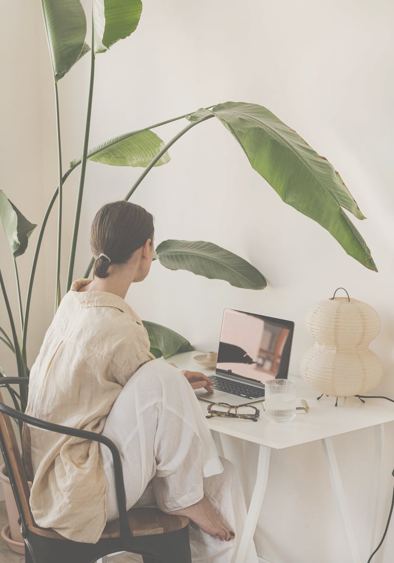 A woman in beige shirt and white trouser sitting at a desk looking into the laptop. Calm scene