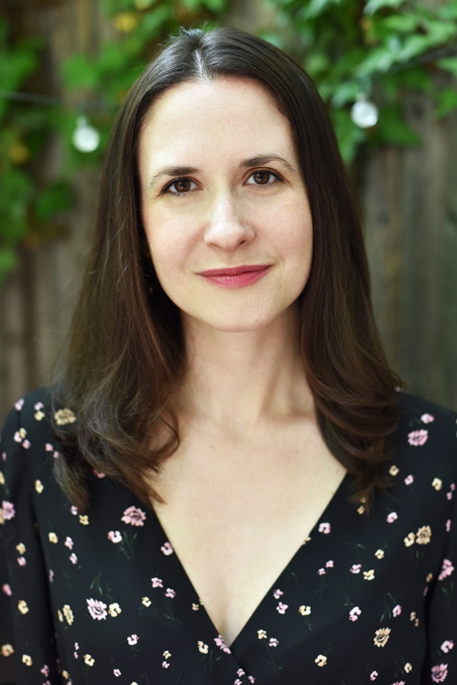 Katie Schorr has shoulder-length dark brown hair and brown eyes. She wears a black floral blouse.