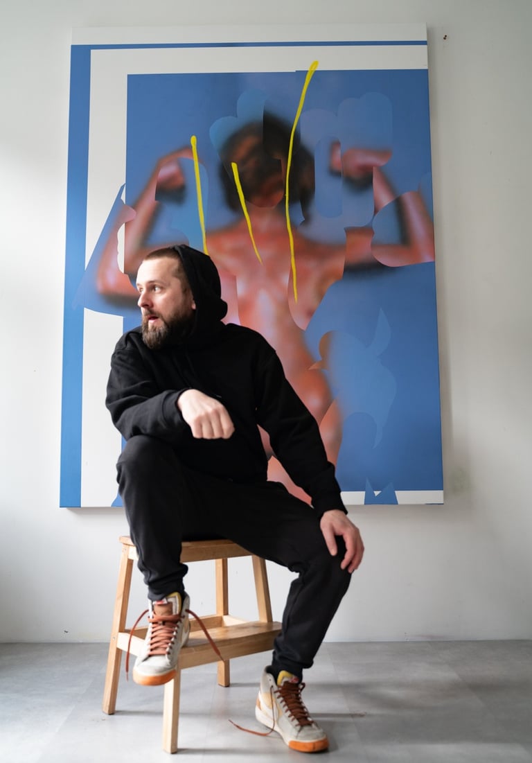 Artist Hadas Patryk in black hoodie sitting on a wooden stool in front of a contemporary painting.
