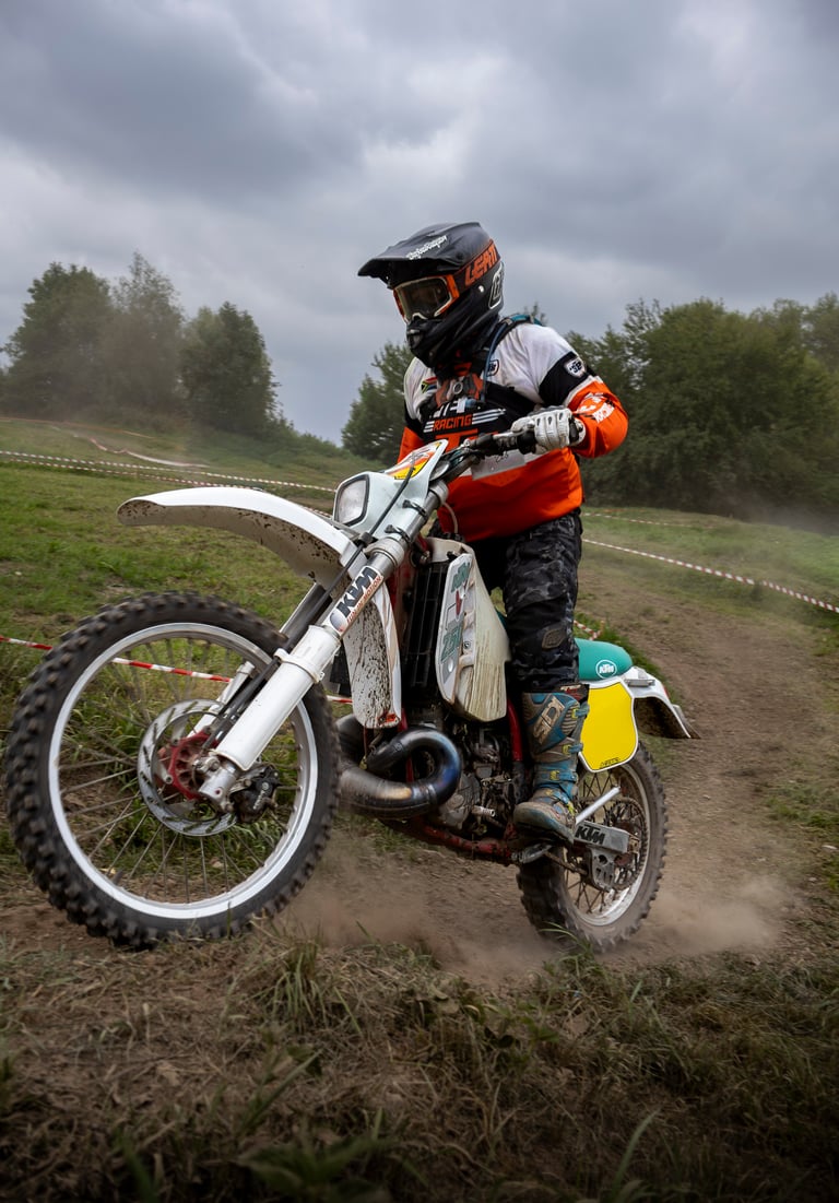 A motocross rider in orange and white gear racing a dirt bike on an off-road track under a cloudy sky.