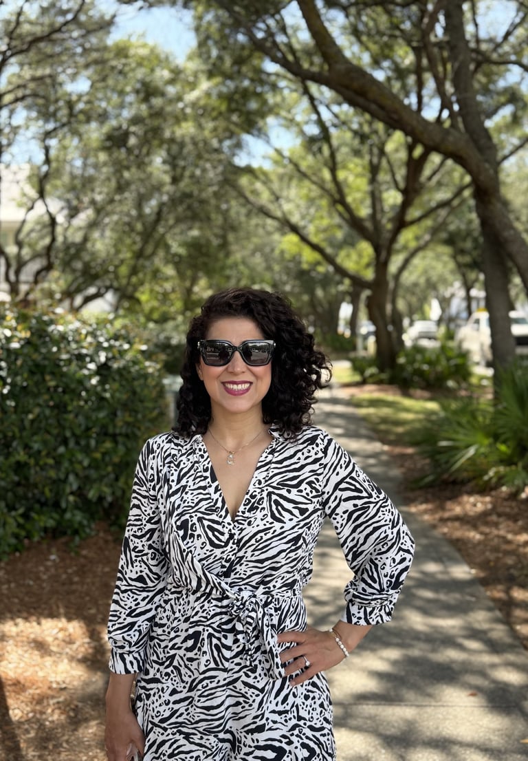 A smiling woman with curly hair wearing a zebra print wrap dress and sunglasses on a sunny tree-lined path.