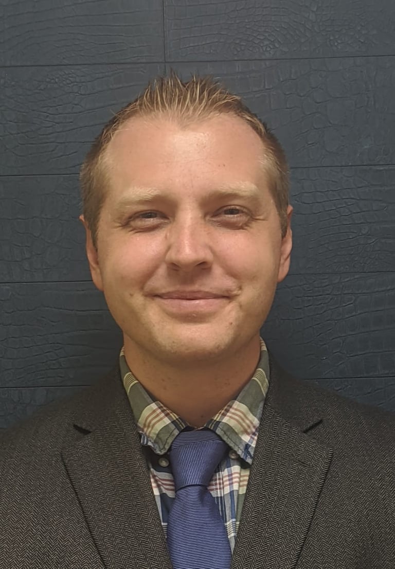 Professional headshot of a smiling man wearing a grey blazer, plaid shirt, and blue tie.