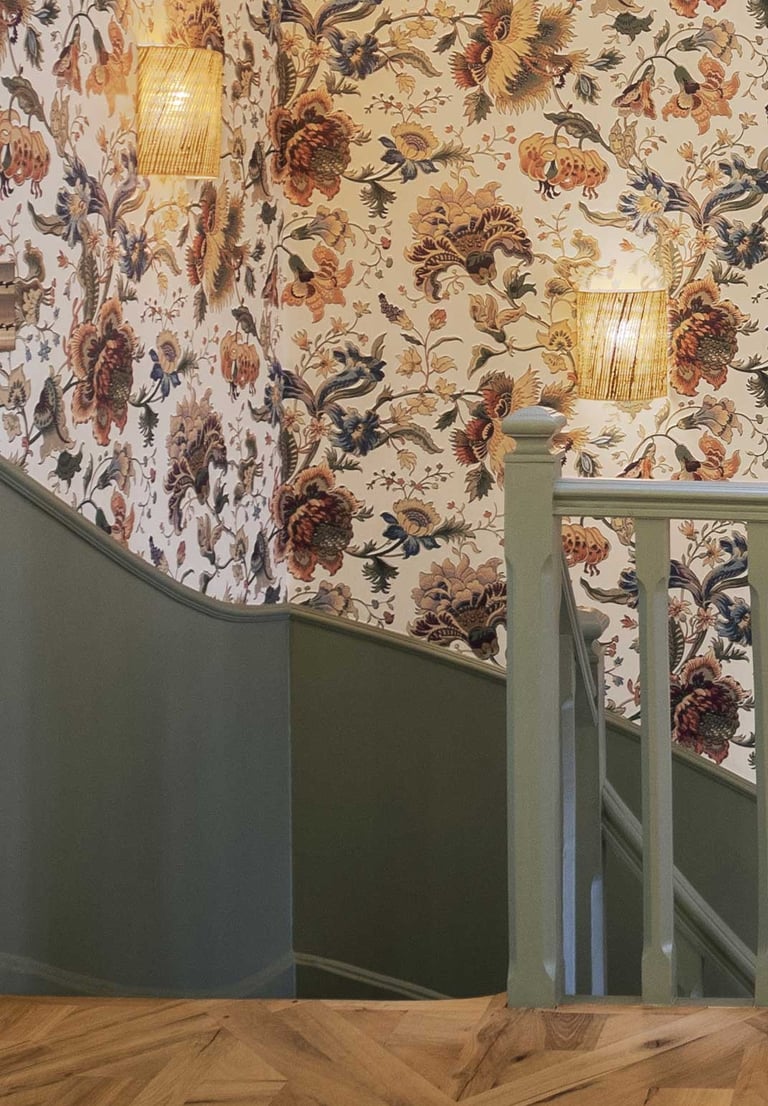Vintage floral wallpaper lines a staircase with sage green wood paneling and woven wall sconces.