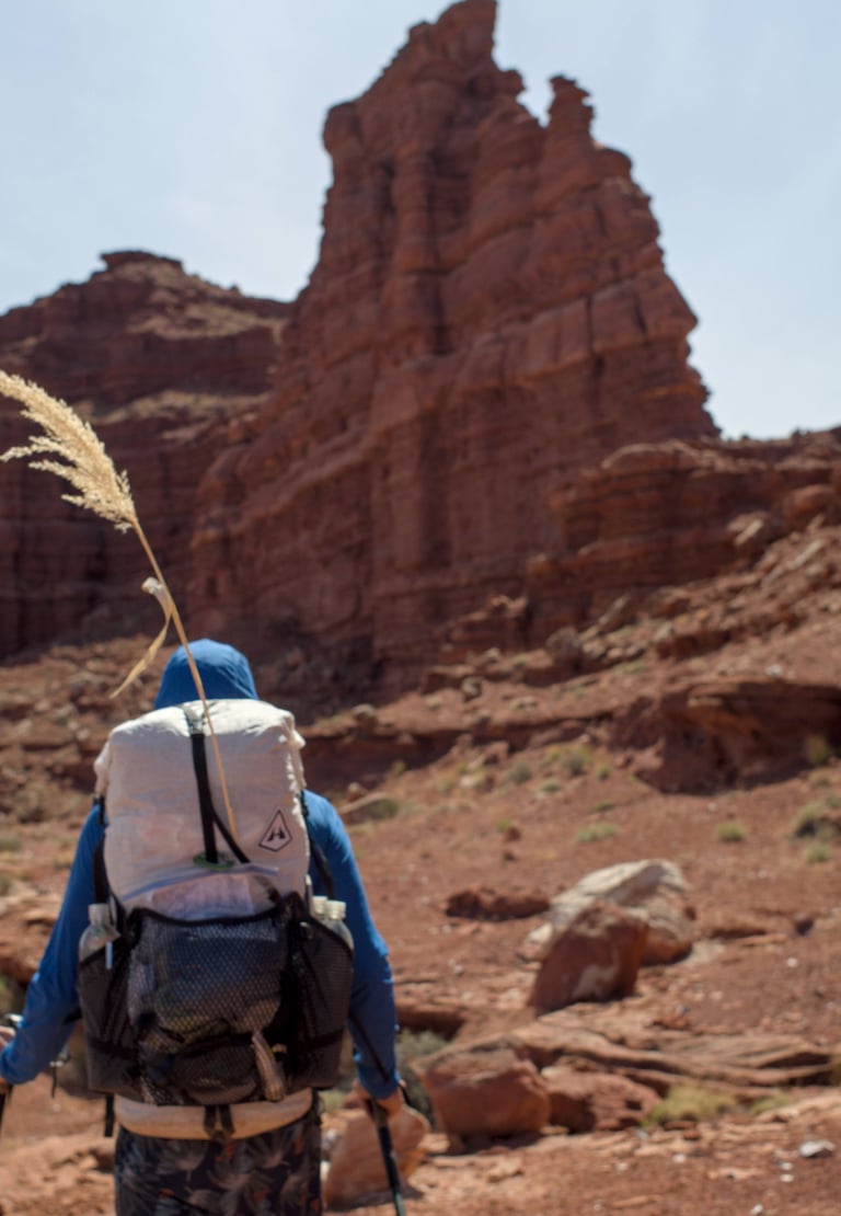 A hiker with a Hyperlite backpack walks toward a massive red rock formation in a desert canyon.