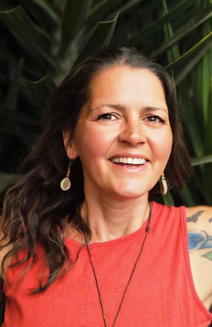 A smiling woman with wavy brown hair wearing a red top and boho earrings in front of green leaves.