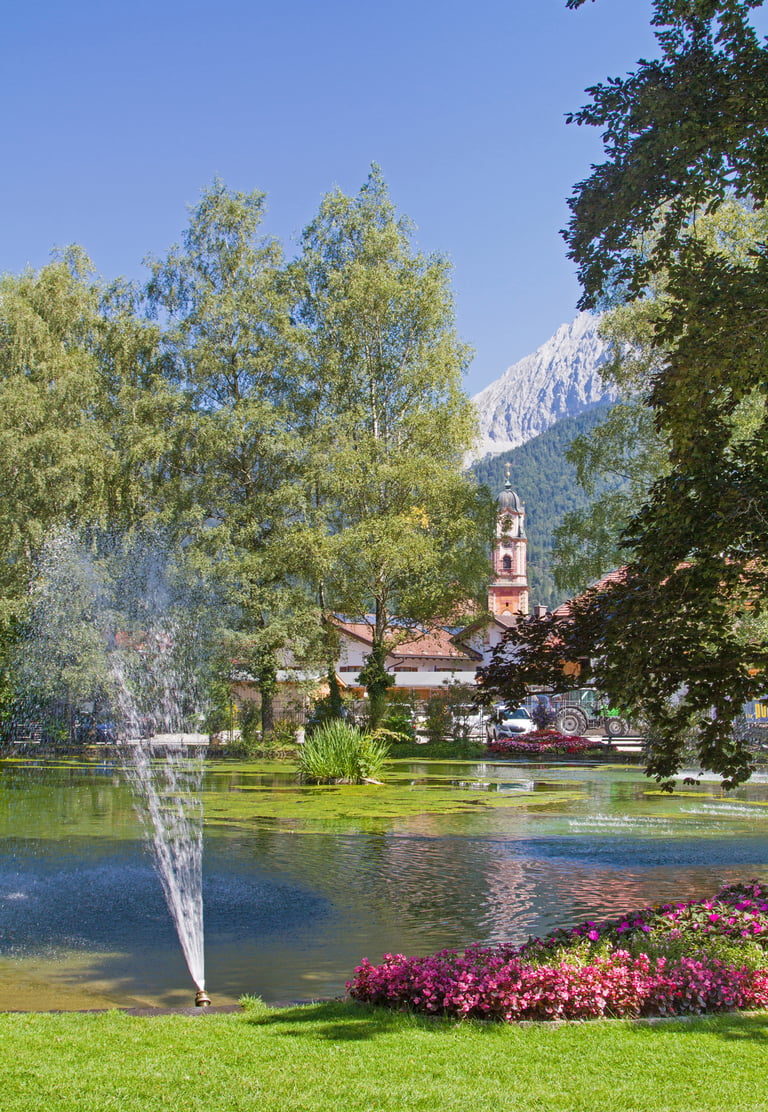 A scenic park in Mittenwald, Germany featuring a pond with a water fountain and the Alps in the distance.