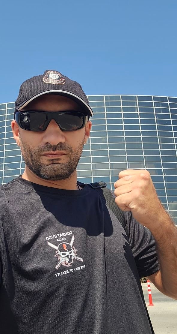 A man wearing a black martial arts shirt, sunglasses, and a baseball cap posing with a clenched fist outdoors.