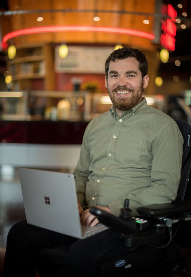Sean wearing a green shirt and holding a silver laptop smiling at the camera in a cafe