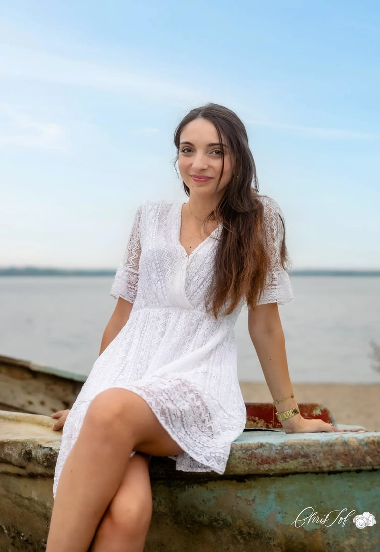 Jeune femme souriante en robe blanche en dentelle assise sur une barque au bord de l'eau à Canet 66.