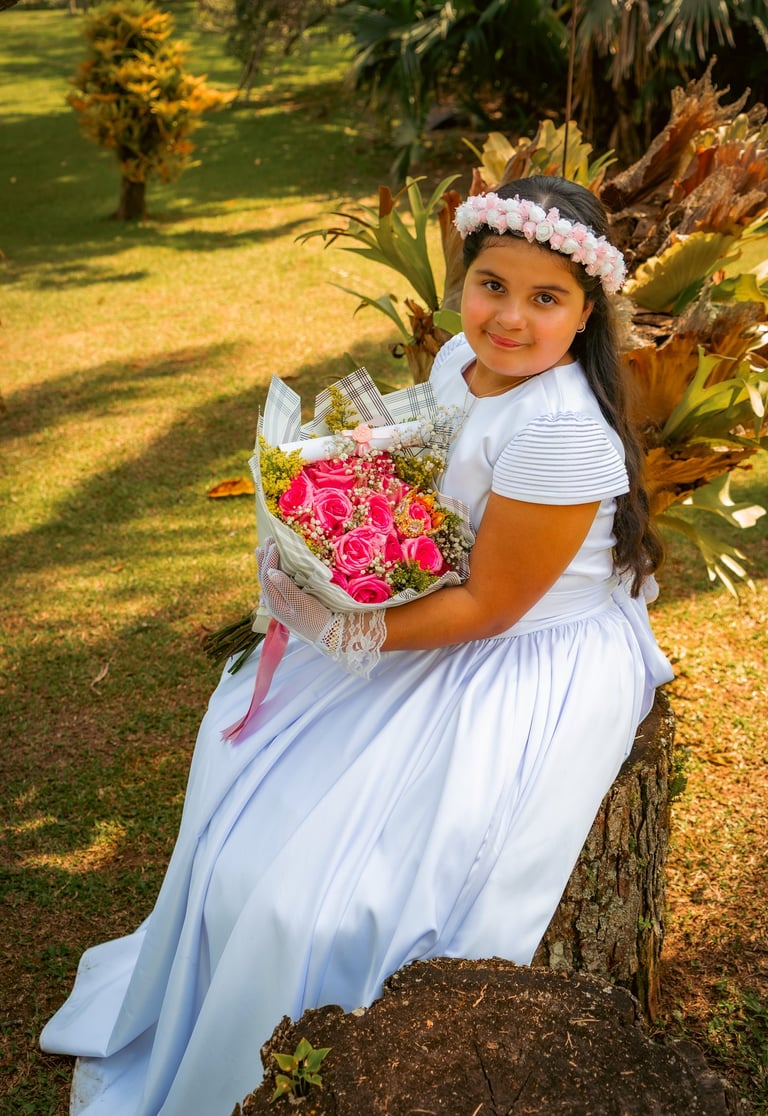 Niña con un vestido de primera comunión blanco sosteniendo un ramo de rosas rosas al aire libre.