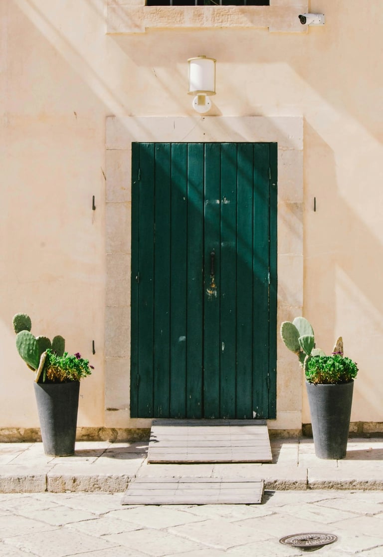 itahome luxury Rustic dark green wooden door on a beige stone wall flanked by potted cactus plants.