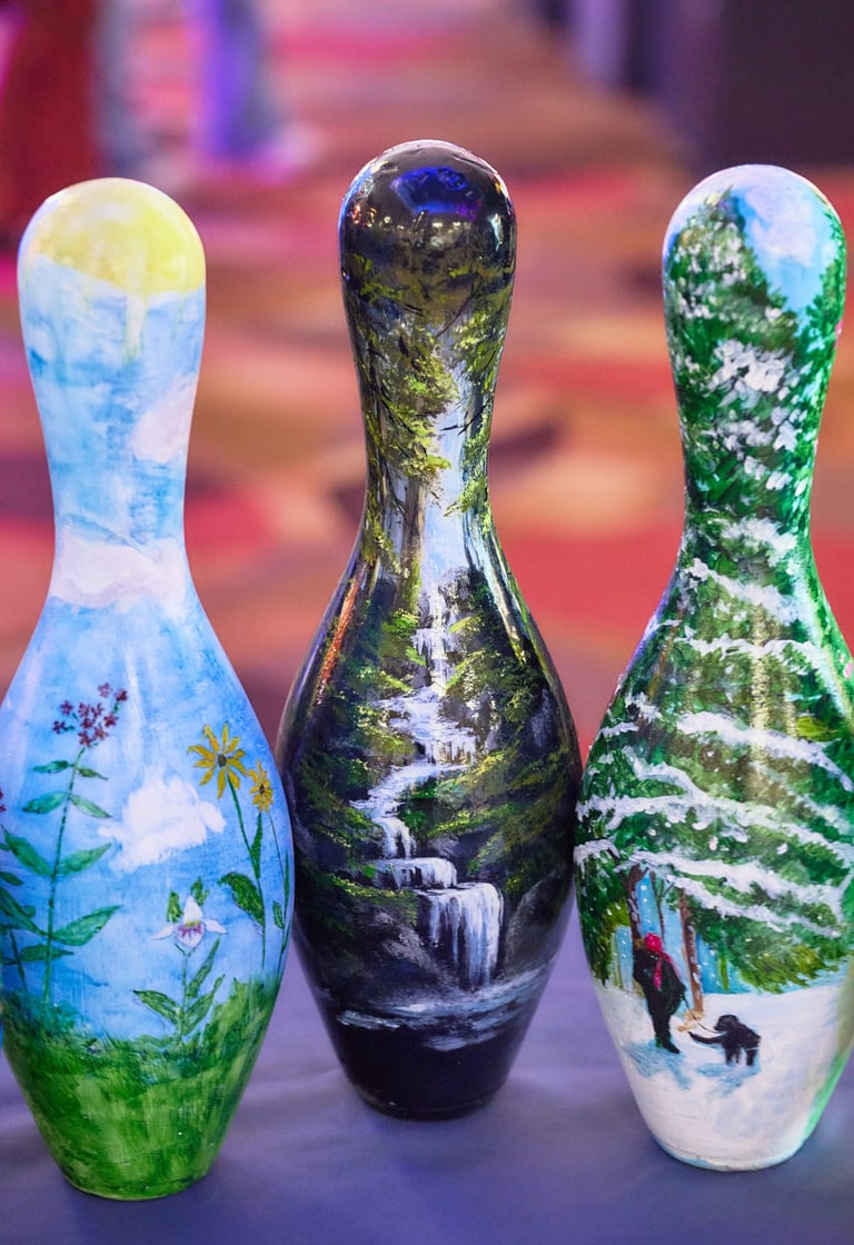 Three hand-painted bowling pins featuring nature landscape art including waterfalls and snowy forests.