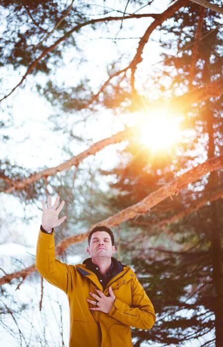 Person in yellow jacket with hand raised, standing in sunlit snowy forest with tall pines.