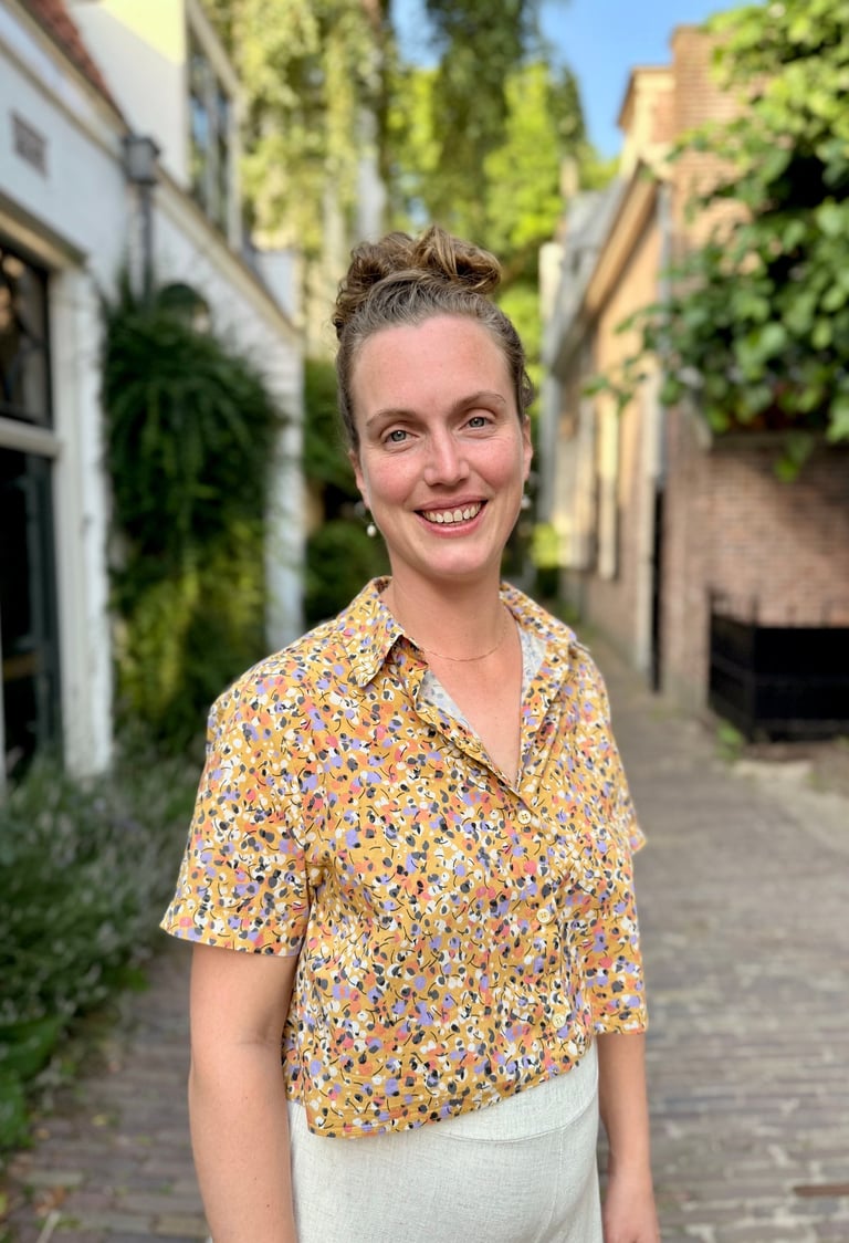 Smiling woman with a hair bun wearing a floral patterned yellow shirt on a cobblestone street.