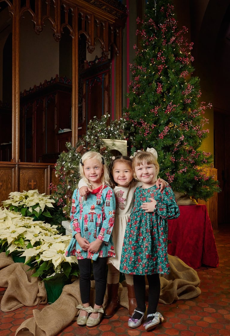 Three young girls in holiday dresses posing by a decorated Christmas tree and white poinsettias.
