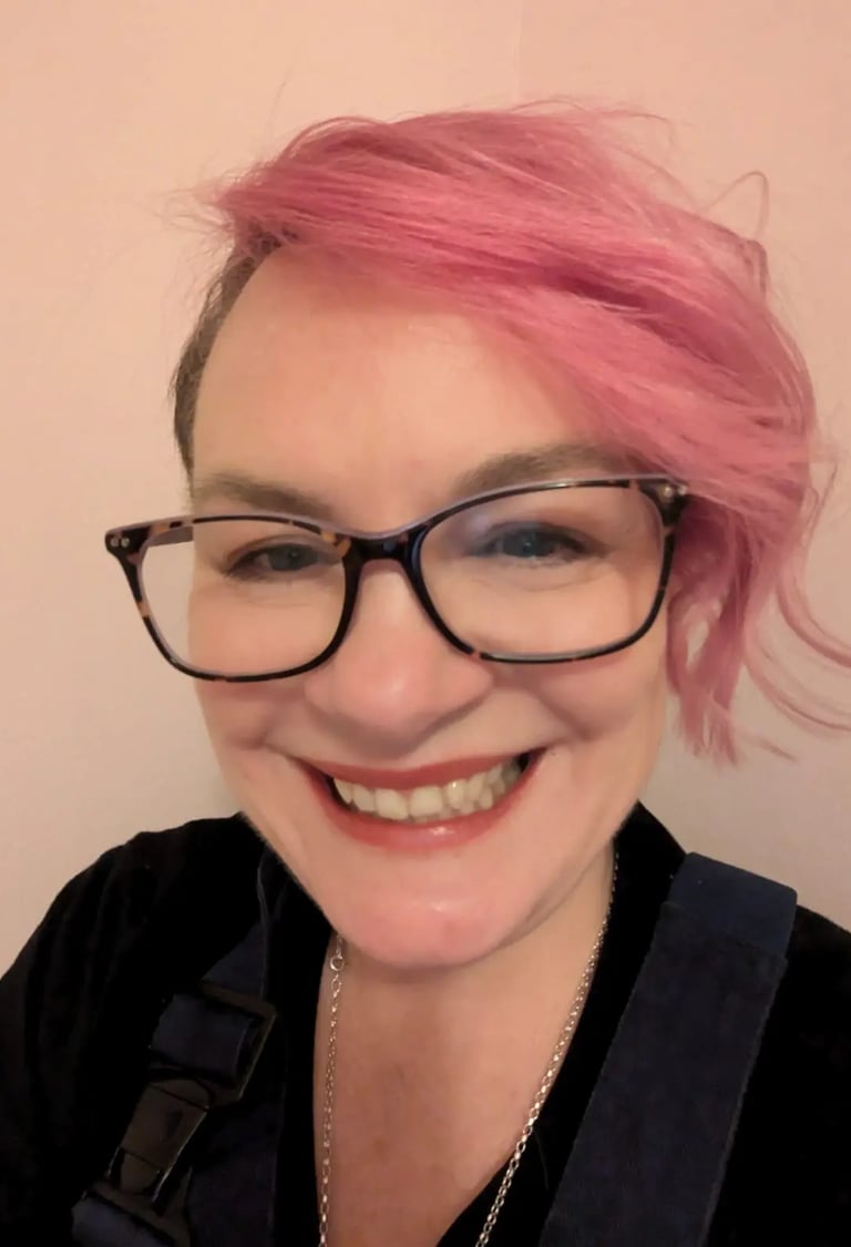 Smiling woman with bright pink hair wearing tortoise shell glasses and black overalls.