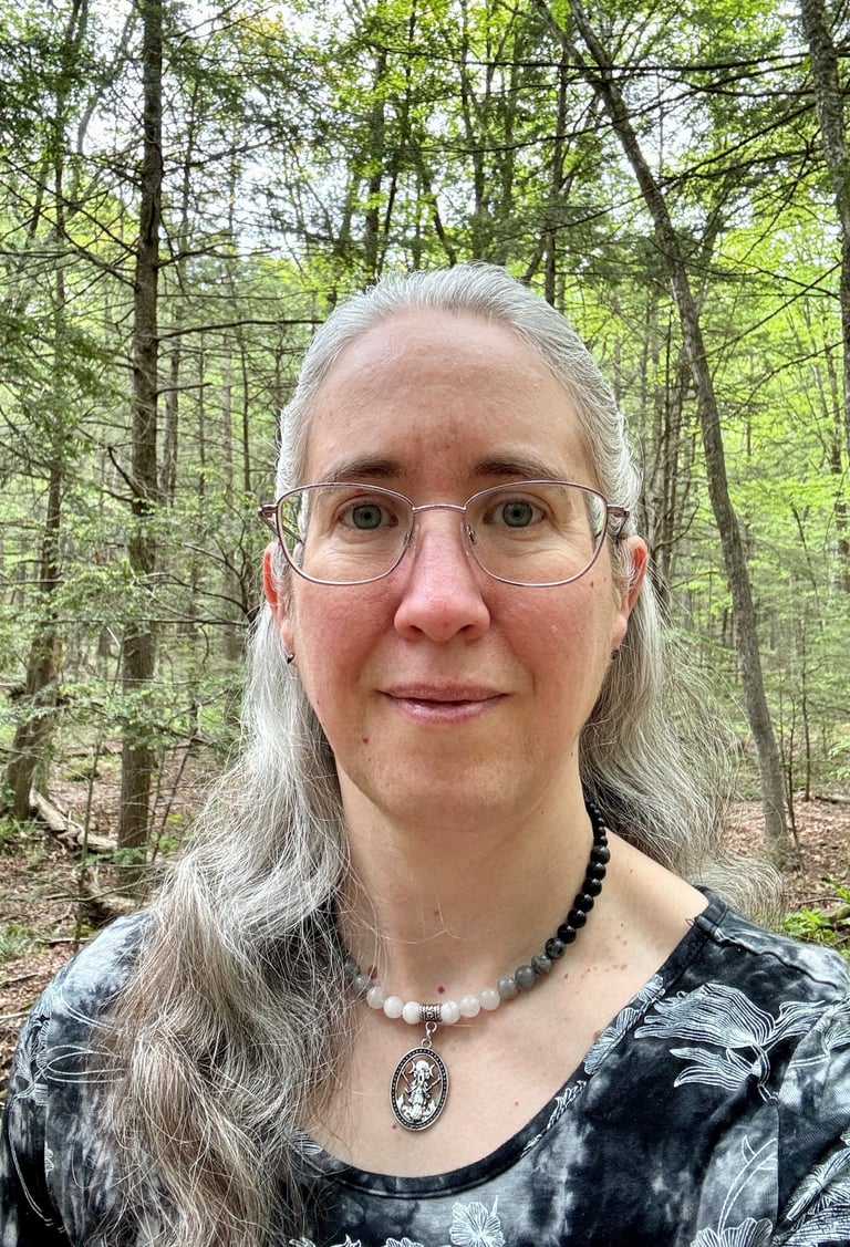 Portrait of a middle-aged white woman with long gray hair and glasses, standing in the woods.