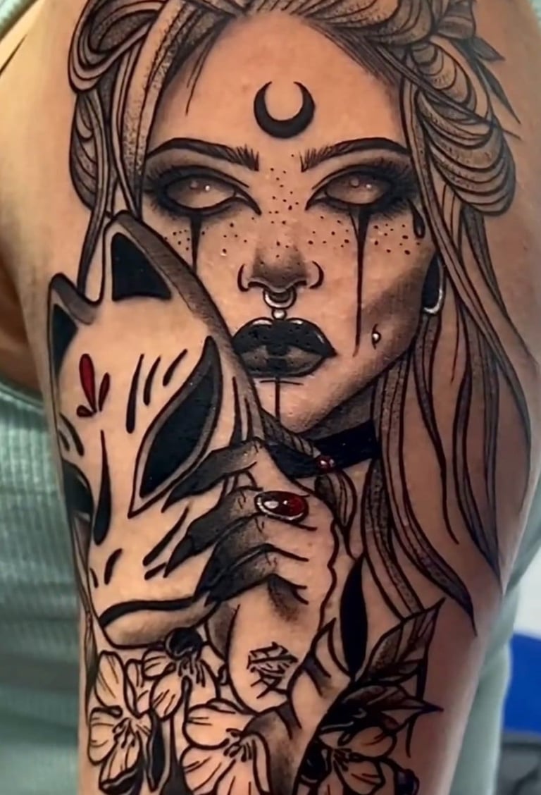 Detailed black and grey ink tattoo on shoulder featuring a gothic woman with a crescent moon and kitsune mask.