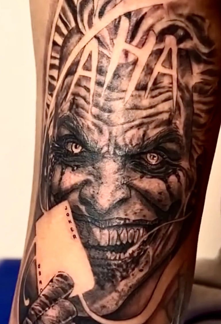 Detailed black and grey Joker tattoo featuring a realistic villainous grin, HA HA text, and a joker playing card.